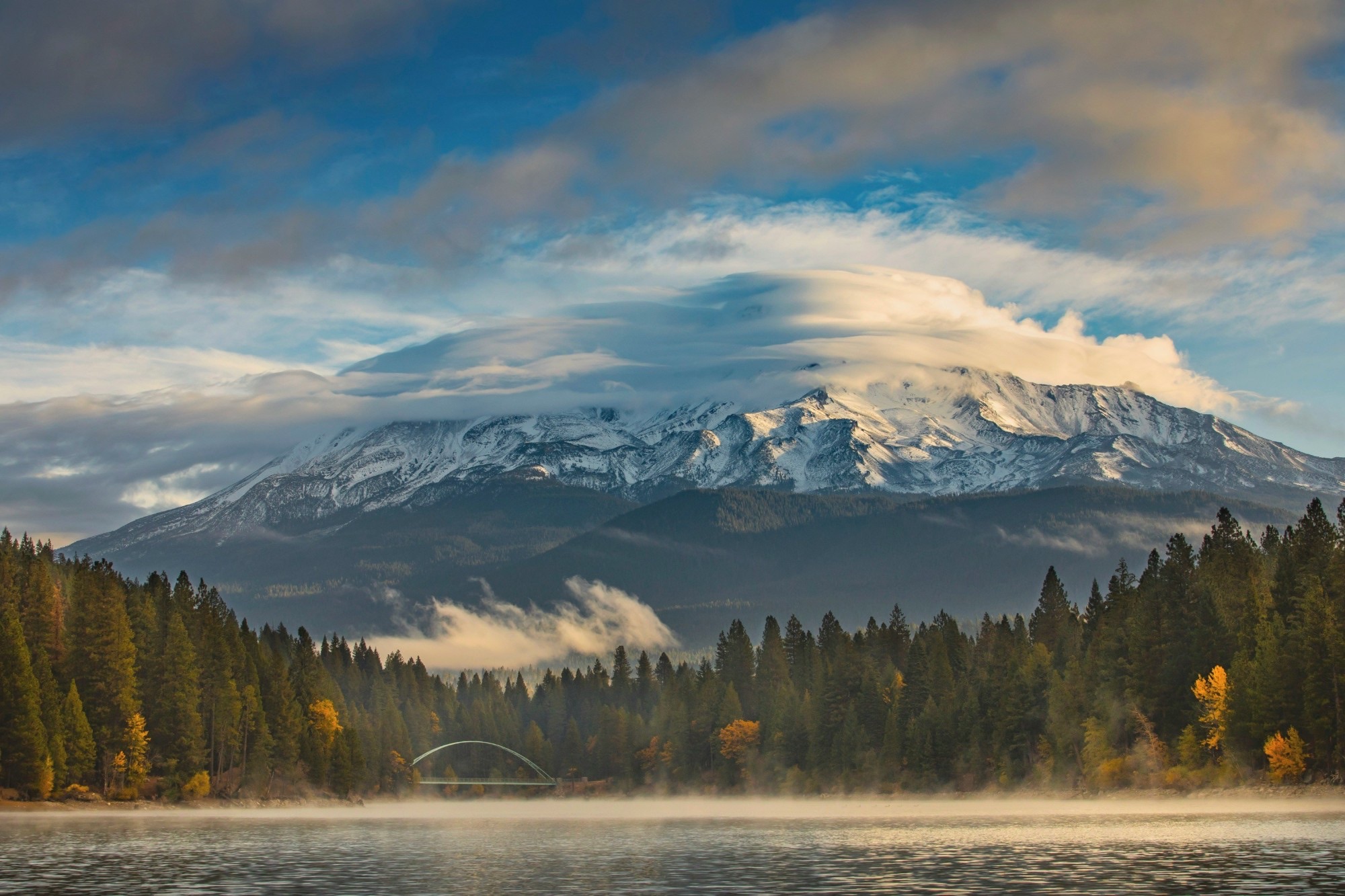 Mount Shasta under lenticular clouds, above a misty lake and forest